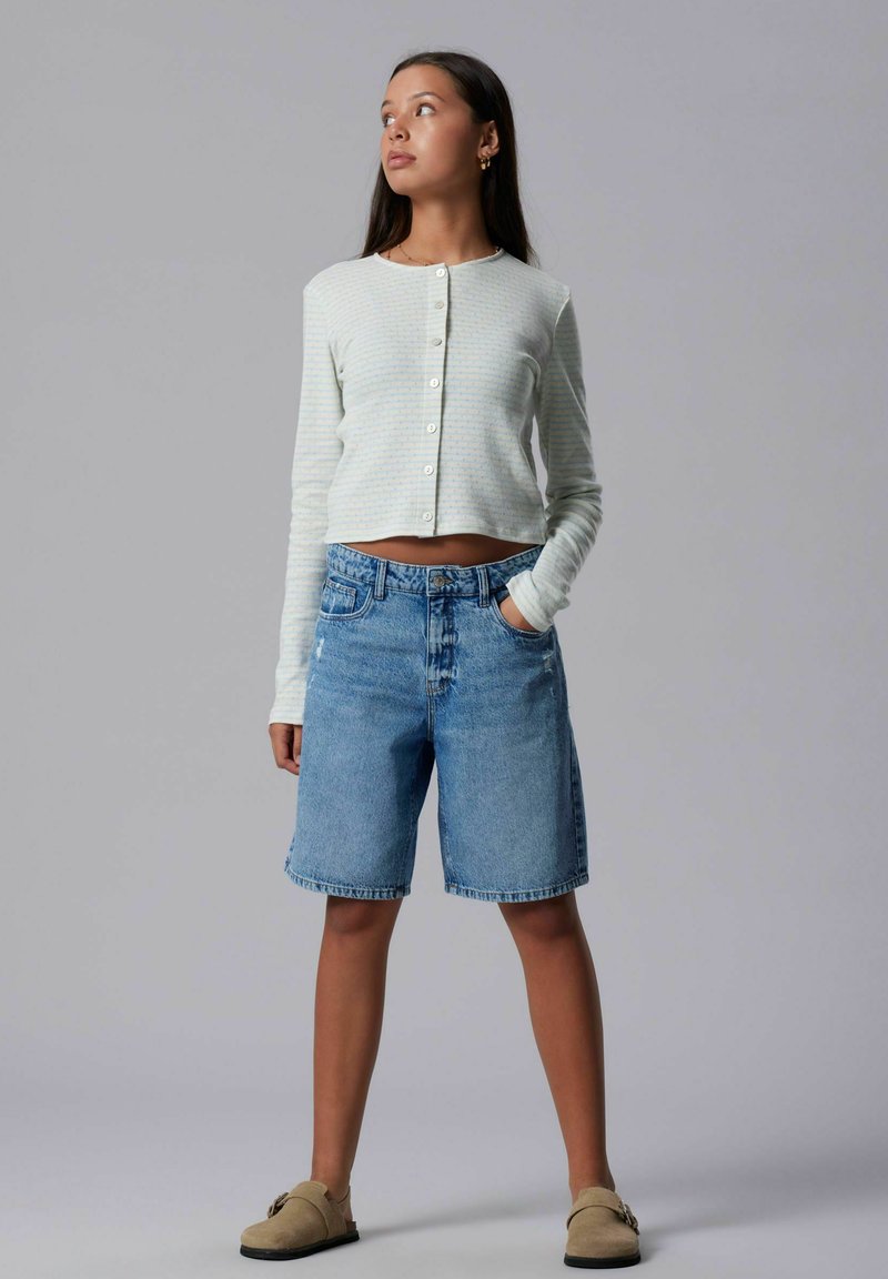 Femme portant un cardigan léger rayé boutonné, un short bermuda en denim bleu et des chaussures beiges à enfiler, debout avec une main dans la poche.