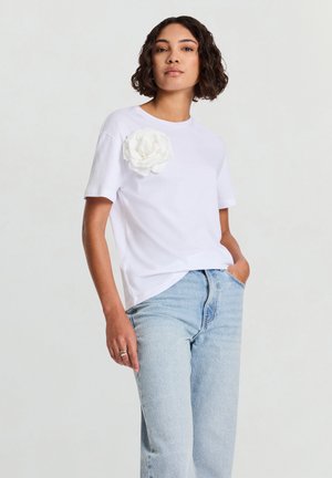 T-shirt bianca in cotone con maniche corte, decorata con un grande fiore di stoffa bianco sul lato sinistro del petto. Indossata con jeans a vita alta di colore azzurro chiaro.