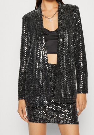Frau trägt einen schwarzen Paillettenblazer und passenden Rock über einem schwarzen Crop-Top und einer silbernen Kettenhalskette.