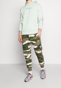Uomo che indossa un felpa leggera verde "Just Do It", pantaloni della tuta Nike mimetici, calzini bianchi e scarpe Nike grigie, in piedi contro un muro bianco.