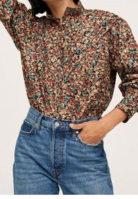 Beżowa koszula z kwiatowym wzorem, z czerwonymi, niebieskimi i zielonymi motywami; zestawiona z jeansami typu high-waist w kolorze niebieskim.