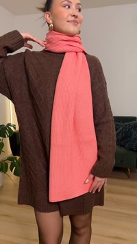 Vestido de malha castanho com textura canelada, combinado com um longo cachecol coral canelado enrolado ao pescoço, num ambiente interior luminoso.