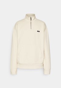 Sudadera de forro polar color crema con cuello alto, media cremallera y un bolsillo canguro frontal. Cuenta con un pequeño logo negro en el pecho.