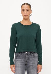 Grønn strikket genser med rund hals og lange ermer, med en avkortet hem og ribbestrikkede mansjetter, kombinert med grå jeans.