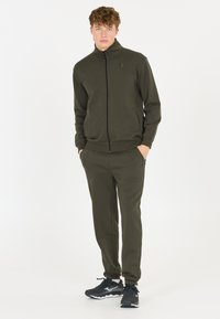 Oliivinvihreä vetoketjuhuppari, jossa on korkea kaulus ja sivutaskut, yhdistettynä samansävyisiin kapeisiin housuihin, joita korostavat mustat urheilukengät.