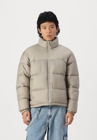 Light gray padded jacket with a high collar and front zipper, featuring horizontal quilted sections and elastic cuffs. Worn over blue jeans.