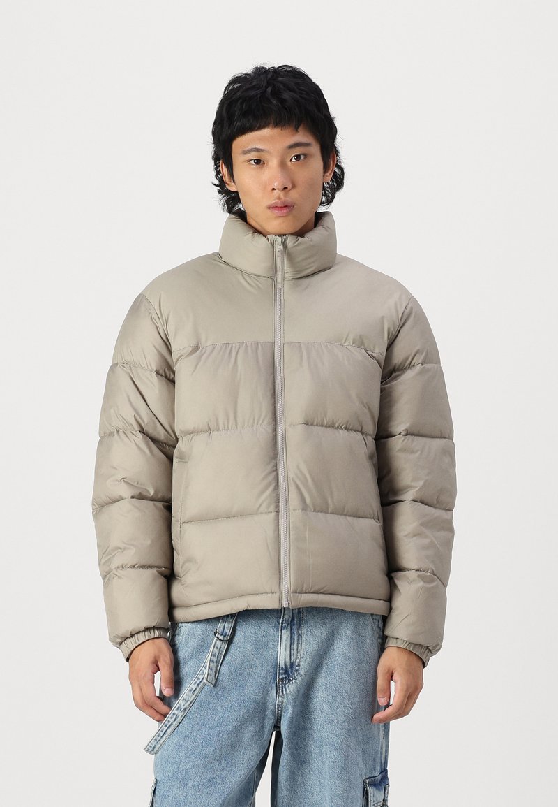 Light gray padded jacket with a high collar and front zipper, featuring horizontal quilted sections and elastic cuffs. Worn over blue jeans.