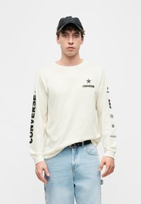 Νέος άντρας που φοράει μαύρο καπέλο, λευκό μακρυμάνικο πουκάμισο Converse με λογότυπα και ανοιχτό μπλε τζιν, στέκεται μπροστά σε απλό φόντο.