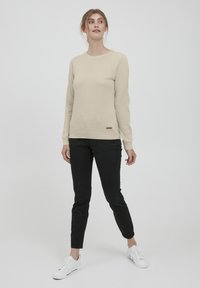 Beige tekstitetu neule, jossa on pyöreä pääntie ja ribbireunat, yhdistettynä slim fit -mustiin farkkuihin ja valkoisiin tenniskenkiin. Hillitty logo-yksityiskohta lantiolla.