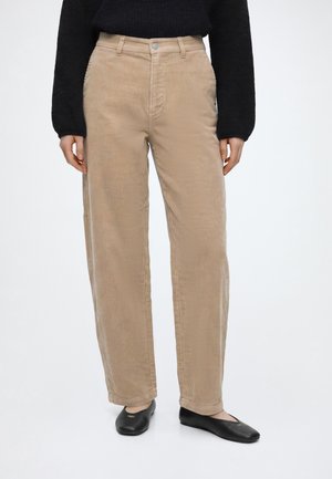 Beige Cordhosen getragen mit einem schwarzen Pullover und schwarzen flachen Schuhen vor weißem Hintergrund, Vorderansicht von der Taille bis zu den Füßen.