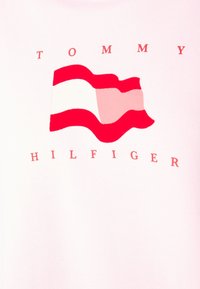 T-shirt rosa chiaro con un design di una bandiera a onde rossa e bianca; "TOMMY HILFIGER" in testo rosso sotto il grafico. Materiale morbido, vestibilità comoda.