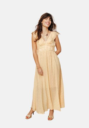 Jeune femme souriante dans une robe maxi sans manches couleur pêche claire avec des volants, portant des sandales dorées et un collier en forme de cœur.