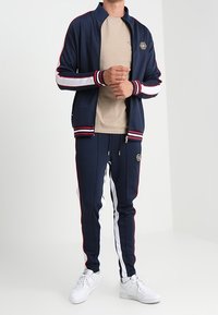 Chándal de color marino con rayas rojas y blancas, que incluye una chaqueta con cremallera y pantalones ajustados. Tejido suave con acentos acanalados y detalle de logo.