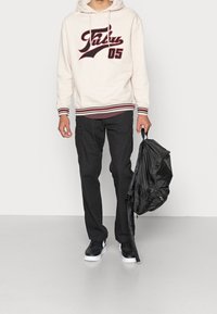 Beige hoodie met donkerrode letters en gestreepte manchetten, gecombineerd met zwarte cargo-broeken. Zwarte rugzak in de rechterhand, witte sneakers.