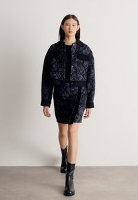 Femme marchant à l'intérieur vêtue d'une veste à motif sombre, d'une jupe assortie, d'un haut noir et de bottines noires, devant un mur uni.