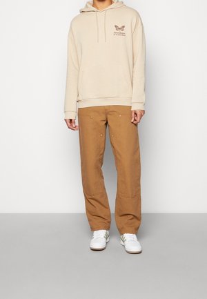 Felpa beige con grafica di farfalla e testo sul petto, abbinata a pantaloni marroni larghi e scarpe da ginnastica bianche con strisce dorate.