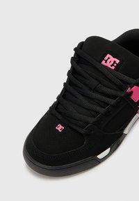 Black sneaker with pink accents, made of suede and synthetic material. Features round laces, perforated toe area, and DC logo on the tongue.