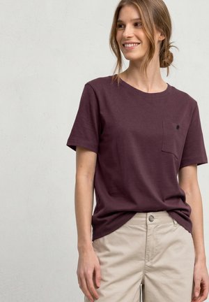 Jeune femme souriante, portant un t-shirt violet foncé à manches courtes avec une poche poitrine, et un pantalon beige, debout devant un mur clair uni.