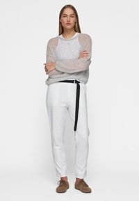 Hellgrauer, transparenter Pullover mit langen Ärmeln, kombiniert mit hellgrauen, engen Hosen und einem schwarzen Gürtel. Braune, strukturierte Schuhe runden das Outfit ab.