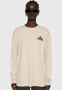 Beige långärmad t-shirt i texturerat tyg, med en grå triangulär logotyp med text på vänstra bröstet.