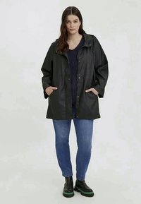 Manteau noir imperméable avec capuche, deux poches latérales, boutons-pression, coupe ample, porté par-dessus un pull sombre et un jean bleu. Bottes noires avec semelles vertes.
