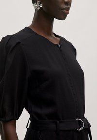 Zwarte jumpsuit met een ronde halslijn, korte pofmouwen, sluiting met rits aan de voorkant, en een bijpassende riem met een zilveren ringgesp.