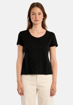 Giovane donna con lunghi capelli castani che indossa una maglietta nera a maniche corte con piccoli pois e pantaloni chiari, in piedi davanti a uno sfondo neutro.