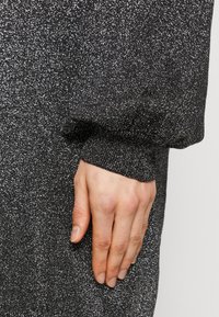 Tessuto a maglia nero glitterato con puntini argento, che mostra un design con maniche a polsino e una mano parzialmente visibile appoggiata sul tessuto.
