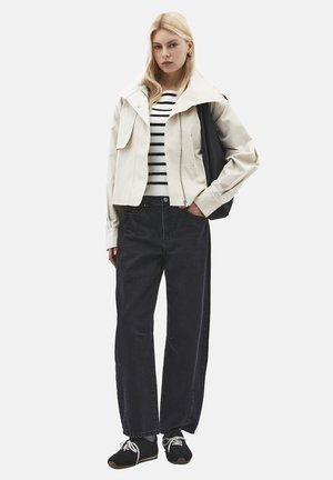 Frau mit langen blonden Haaren, die eine cremefarbene Jacke, einen gestreiften Pullover, lockere schwarze Hosen, schwarze Sneakers trägt und eine schwarze Umhängetasche bei sich hat.