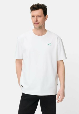 Mand med kort hår iført en enkel hvid T-shirt med en lille grøn logo på brystet og sorte bukser, stående foran en hvid baggrund.