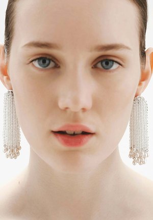 Joven con piel pálida y ojos azules que lleva pendientes largos de cuentas con perlas en los extremos, mirando hacia adelante con expresión neutra.