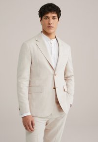 Helles beiges Leinenanzug mit einreihigem Sakko, zwei vorderen Taschen und passender Hose. Weißes Hemd mit Kragen ist darunter sichtbar.