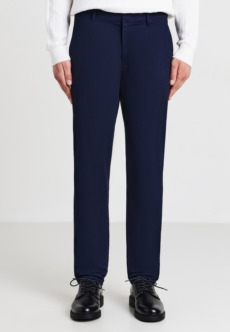 Homme portant un pantalon bleu foncé, des chaussures noires à lacets et un pull à manches longues à côtes blanc se tenant sur un fond clair.