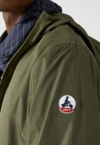 Veste légère vert olive avec une texture lisse, présentant un badge logo distinctif JOTT sur l'épaule et une fermeture éclair à l'avant.
