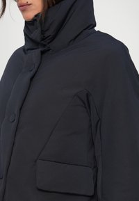 Zwarte gewatteerde gewatteerde jas met capuchon, gemaakt van geborduurd materiaal. Kenmerkt zich door grote knopen, een driehoekige zak en een hoge, gestructureerde kraag.