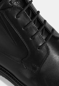 Gros plan sur une chaussure habillée en cuir noir montrant les détails de couture et de lacets sur une surface lisse et polie.