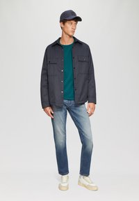 Marineblaue Jacke mit Knöpfen über einem grünen Hemd, kombiniert mit hellblauen Jeans und weißen Sneakers. Das Model trägt eine passende marineblaue Mütze.