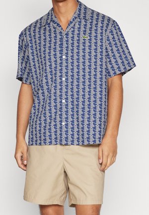 Mężczyzna w niebieskiej koszuli z krótkim rękawem w geometryczny wzór oraz beżowych szortach, stojący na jednolitym tle.