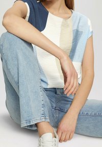 Kortærmet top med tekstureret stof, med blå, creme og hvide abstrakte mønstre, kombineret med lyseblå jeans og hvide sneakers.