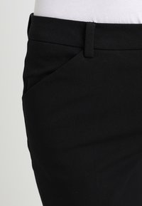 Pantalons en tissu noir avec une texture lisse, présentant une poche coupée en diagonale et un design ajusté. La ceinture est plate et discrète.