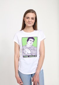 Bílý tričko s potiskem znázorňujícím postavu s tmavými vlasy. Design obsahuje zelené a fialové akcenty a text "THE HULK".