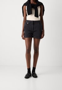 Pantalones cortos negros de corte entallado, con bolsillos frontales y cierre de botón visible. Combinados con una camiseta de tirantes de color claro y un suéter negro.