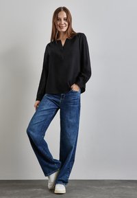 Blouse noire à manches longues avec un décolleté en V, associée à un jean en denim bleu ample et des baskets blanches, le tout sur un fond neutre.
