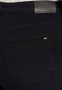Bolsillo trasero de unos vaqueros negros de Tommy Hilfiger con un pequeño logo de bandera rojo, blanco y azul y una etiqueta de cuero negro de la marca encima de la cinturilla.
