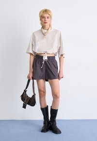 Top corto a righe beige chiaro con cordoncino. Pantaloni corti a righe scure, stivali neri al ginocchio e una piccola borsa marrone, tutti esposti in uno studio.