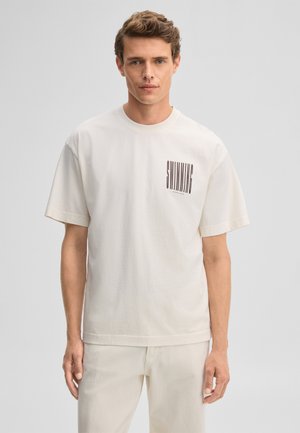 Mann mit kurzen lockigen Haaren, der ein übergroßes off-white T-Shirt und Hosen trägt, stehend vor einem schlichten hellgrauen Hintergrund.