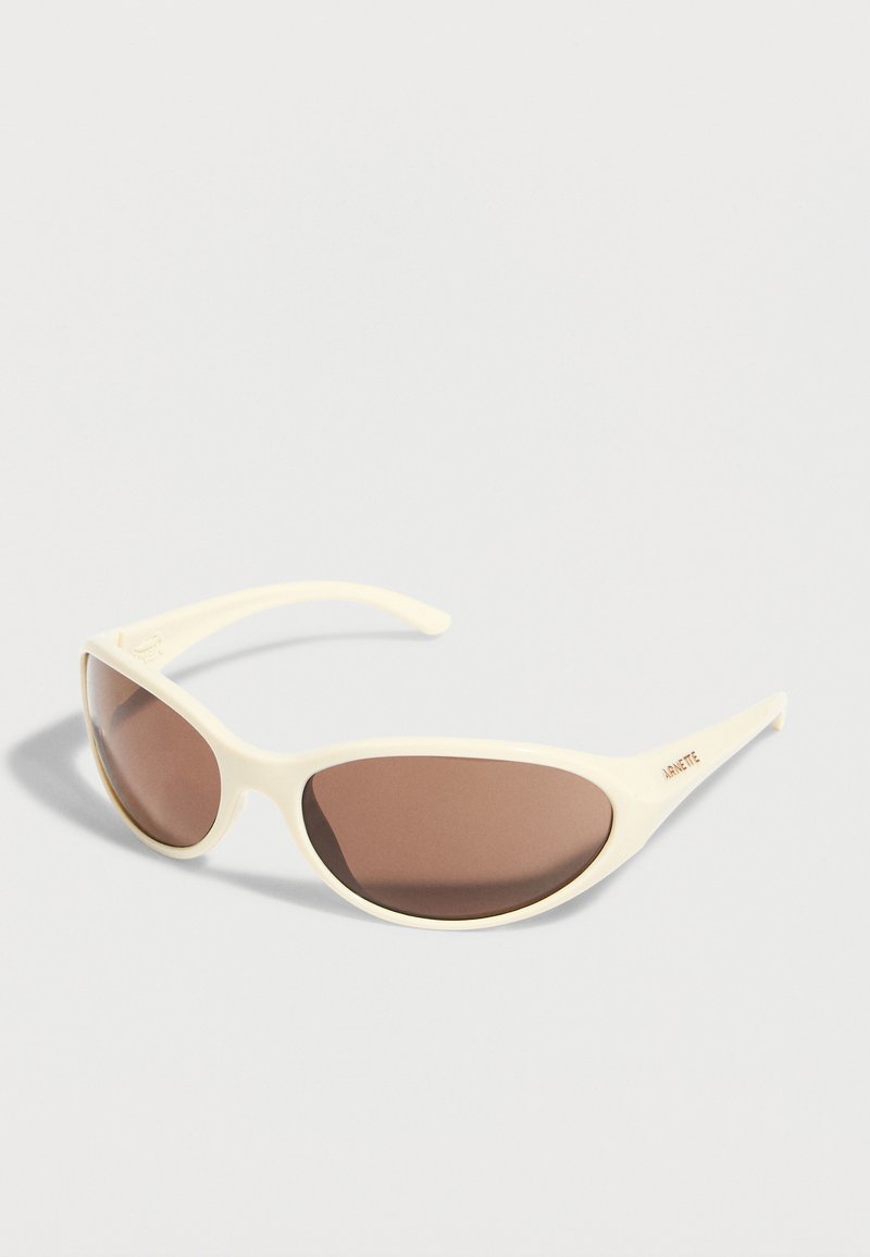 Γυαλιά ηλίου σε κρεμ απόχρωση με καφέ φακούς και το λογότυπο "ARNETTE" στο πλάι, τοποθετημένα πάνω σε λευκή επιφάνεια.
