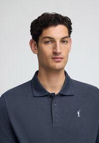 Granatowa koszulka polo z fakturowanej tkaniny, z trzema guzikami z przodu oraz małym jasnoniebieskim logo na lewej piersi.