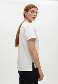 White short-sleeve T-shirt with a relaxed fit, featuring side slits and a smooth, soft texture, paired with black pants.