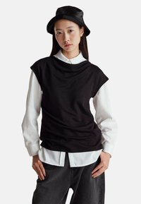 Černý bezramínkový top vrstvený přes bílou košili s knoflíky, doplněný tmavými džíny. Modelka má na sobě černý klobouk. Hladké textury, uvolněný střih.